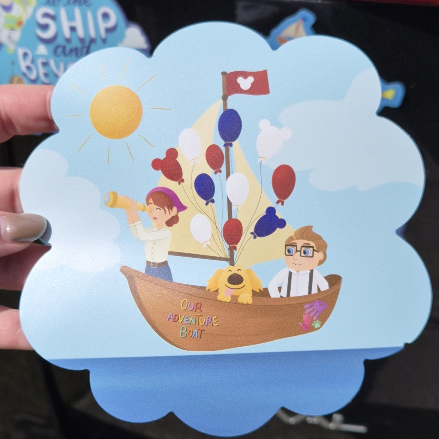 A close-up of a hand holding a custom die-cut Up-themed Disney cruise stateroom door magnet. Features Carl and Russell in a flying hot air balloon boat with the exact text: "Our Adventure Boat". Perfect for cabin decor.