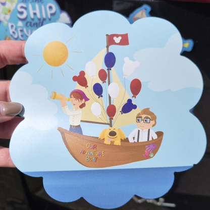 A close-up of a hand holding a custom die-cut Up-themed Disney cruise stateroom door magnet. Features Carl and Russell in a flying hot air balloon boat with the exact text: "Our Adventure Boat". Perfect for cabin decor.