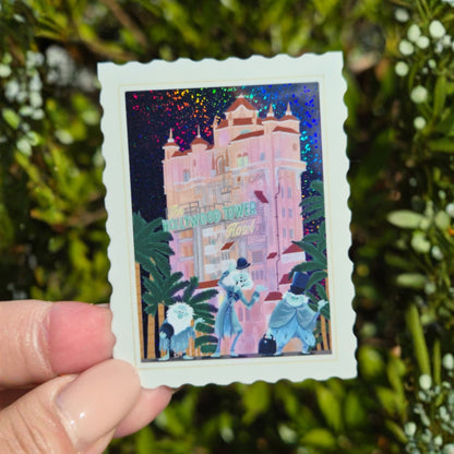 Holographic sticker of a pink building with two characters in front, held against a green leafy background.