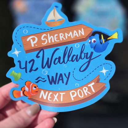 A close-up of a single custom die-cut magnet inspired by Finding Nemo for cruise stateroom doors. A hand holds the magnet, which features Dory and Nemo with prominent integrated text in different fonts: "P. SHERMAN", "42 Wallaby Way", "WAY", "NEXT PORT". The blue magnet includes other illustrated elements like a boat and bubbles. Perfect for adding personality to a Disney cruise cabin door.