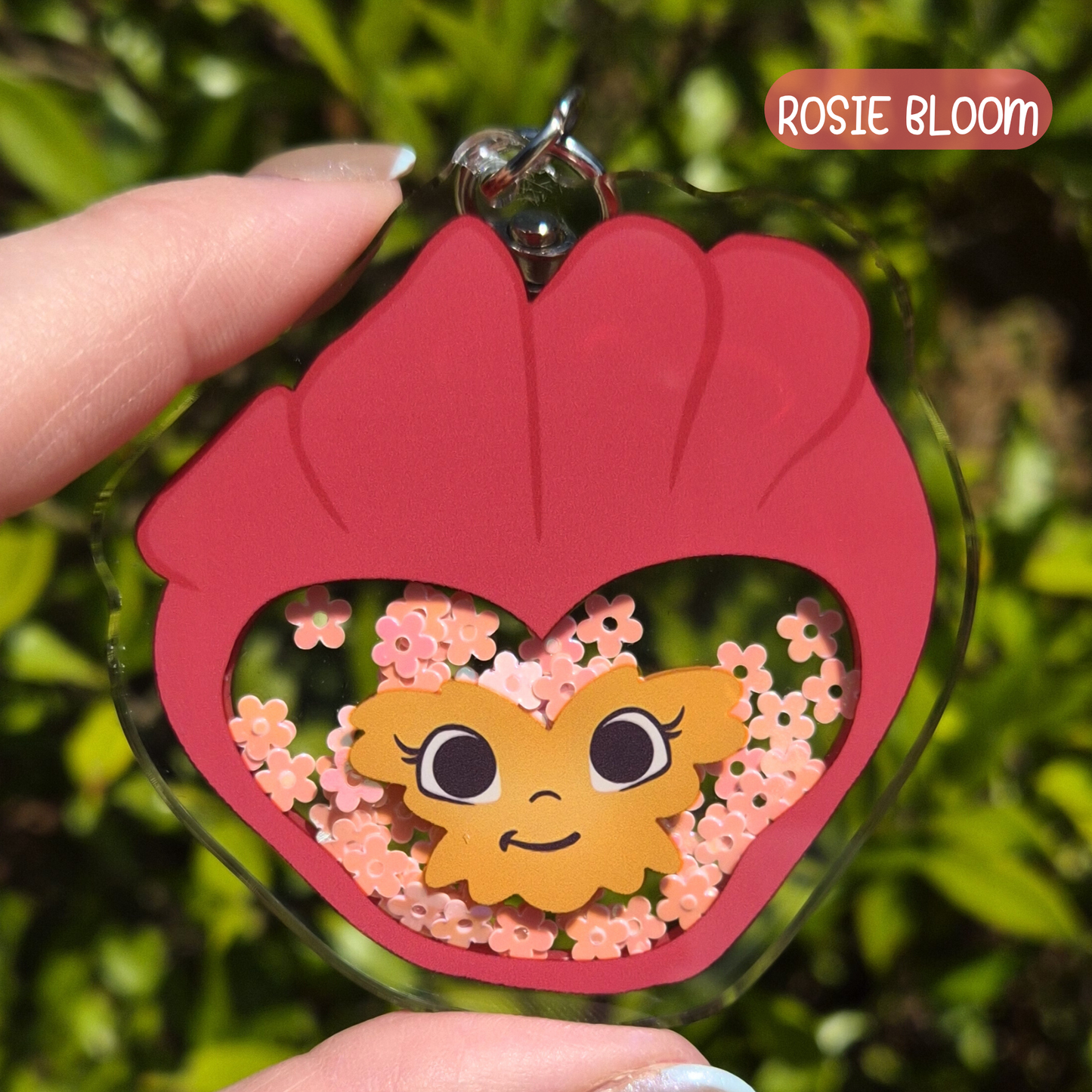Keychain with a cartoon sun design and pink heart frame, held by a hand against a green leafy background.