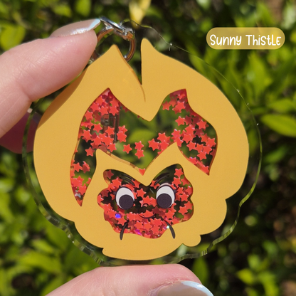 Hand holding a keychain with a cat design against a green leafy background