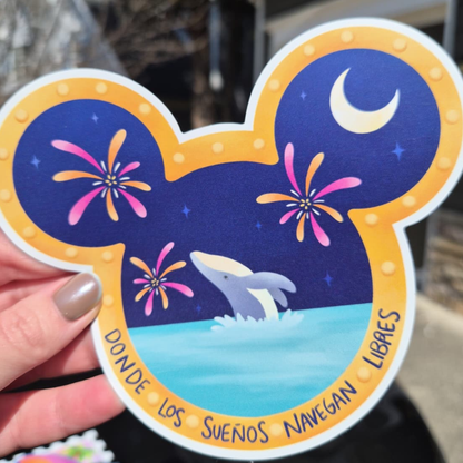 A close-up photo of a hand holding a custom die-cut magnet shaped like Mickey Mouse ears. Inside, a dolphin jumps in front of fireworks at night. Spanish text at the bottom reads: "DONDE LOS SUEÑOS NAVEGAN LIBRES". Perfect for Disney cruise stateroom door decor.