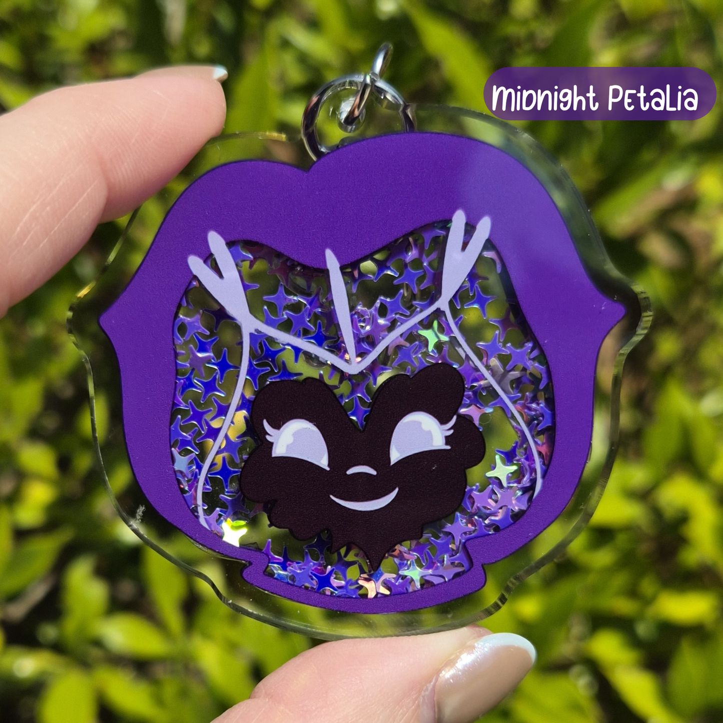 Keychain with a cartoon character inside a purple lip-shaped frame, held by a hand against a green leafy background.