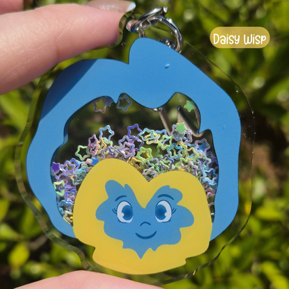 Keychain with a blue and yellow character and star-shaped design, held against a green leafy background.
