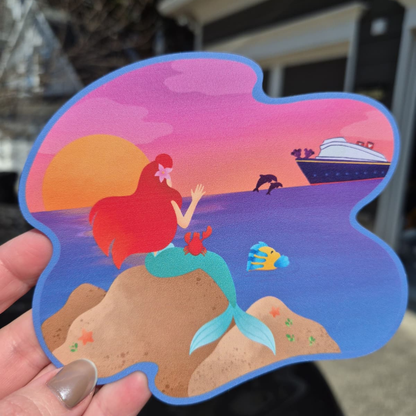 A close-up of a custom die-cut Disney cruise stateroom door magnet held by a hand. The design features Ariel from The Little Mermaid sitting on a rock alongside a crab and a fish, watching a Disney cruise ship at sunset.