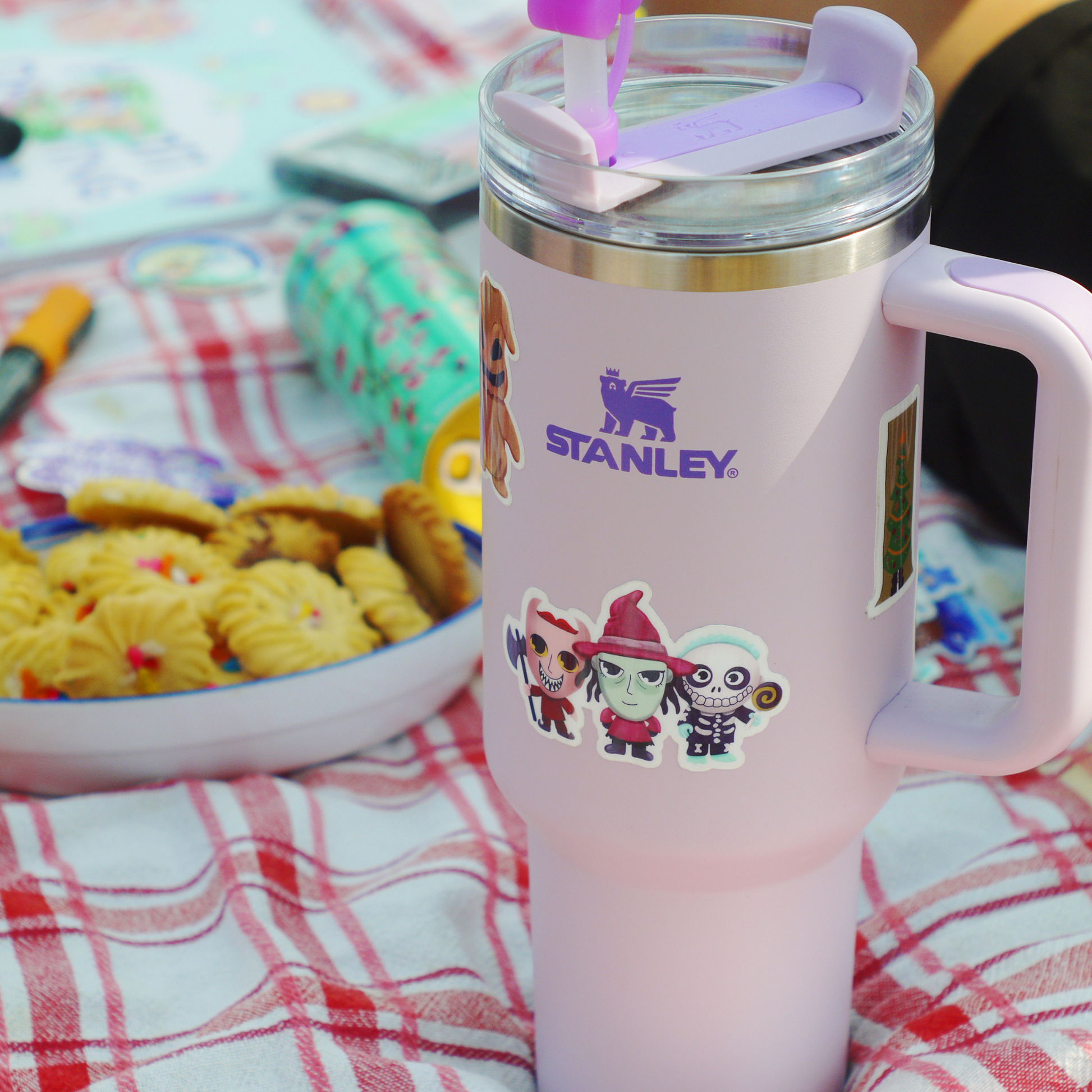 Stanley tumbler with cartoon characters on a checkered tablecloth with cookies and a book.