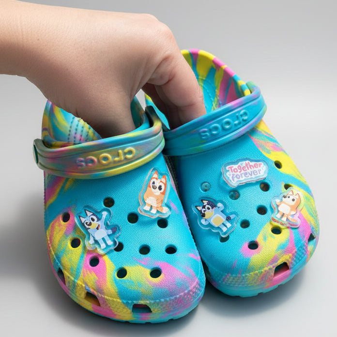 Colorful children's clogs with cartoon characters held by a hand against a garden background.