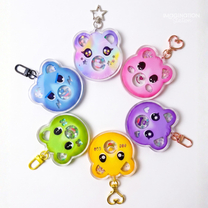 Six colorful bear-shaped keychains arranged on a white background, each with a different pastel color (blue, lavender, pink, green, yellow, purple) and a small heart-shaped charm inside. The keychains have clear plastic edges, cute cartoon eyes, and metal keyrings and clips in silver, rose gold, and gold.