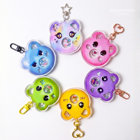 Six colorful bear-shaped keychains arranged on a white background, each with a different pastel color (blue, lavender, pink, green, yellow, purple) and a small heart-shaped charm inside. The keychains have clear plastic edges, cute cartoon eyes, and metal keyrings and clips in silver, rose gold, and gold.