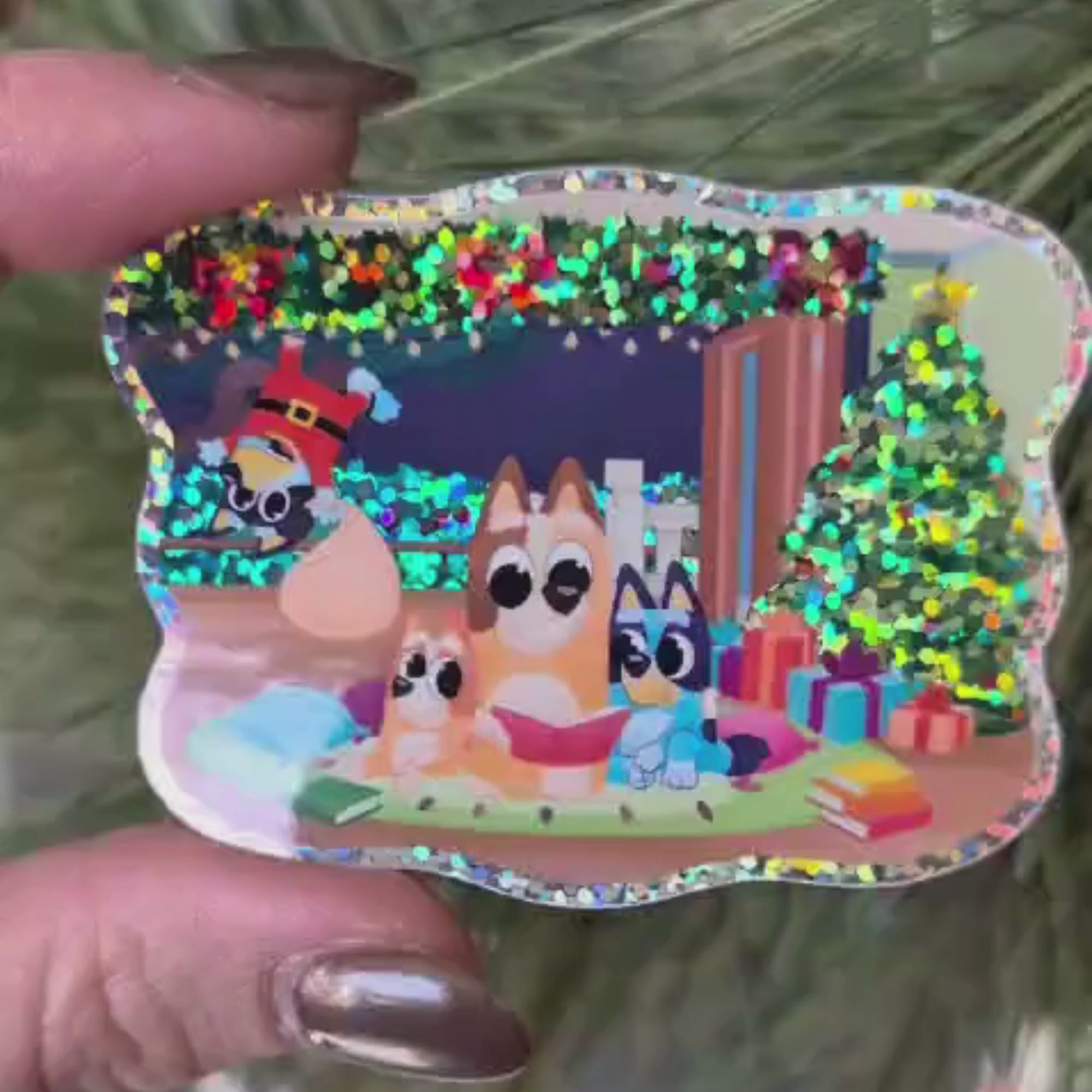 Hand holding a festive Bluey sticker with a sparkling glitter finish, tilting it back and forth to reveal the Christmas overlay effect. Perfect for holiday decoration