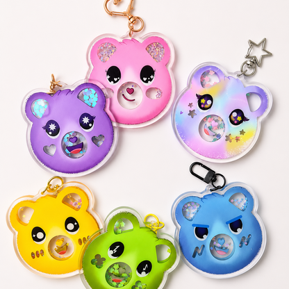 Six colorful bear-shaped keychain pendants arranged on a plain light background. The bears are pink, purple, iridescent blue, yellow, green, and blue, each with glittery ears, embroidered facial details, and metal keychain attachments including heart, star, and clip styles.
