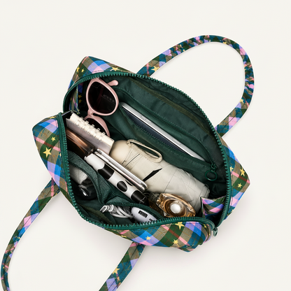 A mini tote bag with a multicolor plaid pattern and star accents, shown open to reveal its dark green interior packed with accessories. Large text at the top reads 'MINI SIZE MAXIMUM STORAGE,' with callout lines pointing to the bag's capacity and noting it can hold an iPhone 17 Pro and a folding umbrella.
