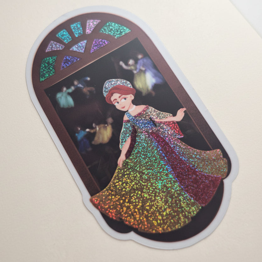 A die-cut sticker featuring an iridescent, glittery illustration of a woman in a flowing multicolor dress, set against a dark background with a stained-glass style arch at the top, shown on a beige surface.