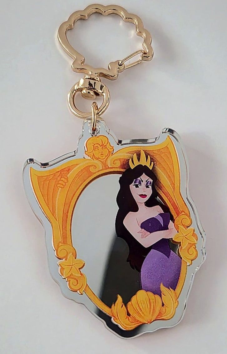 An acrylic keychain featuring a stylized portrait of Ariel from The Little Mermaid inside a gold ornate frame, attached to a gold key ring and clasp, shown on a neutral light gray background.