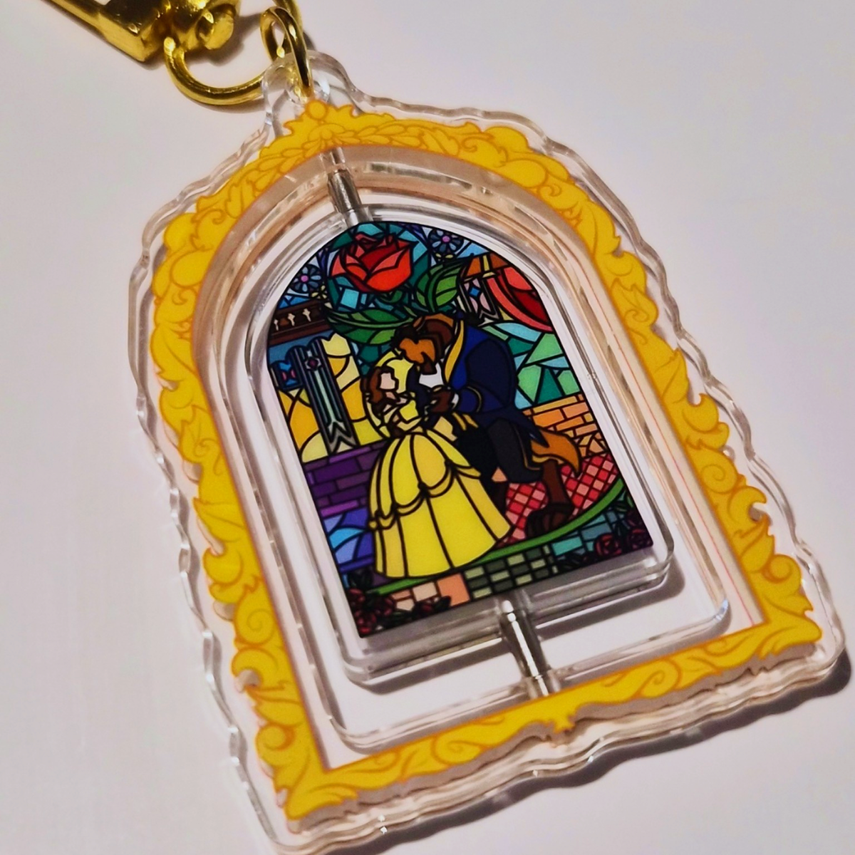 A clear acrylic keychain charm featuring a stylized stained-glass window design with Belle and the Beast from Beauty and the Beast, surrounded by a yellow ornate border and a gold key ring, shown on a plain white background.