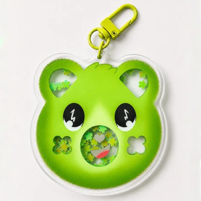 A green bear-shaped keychain or bag charm with a yellow clasp, featuring cartoon eyes, a smiling mouth, and small flower cutouts on a white background.