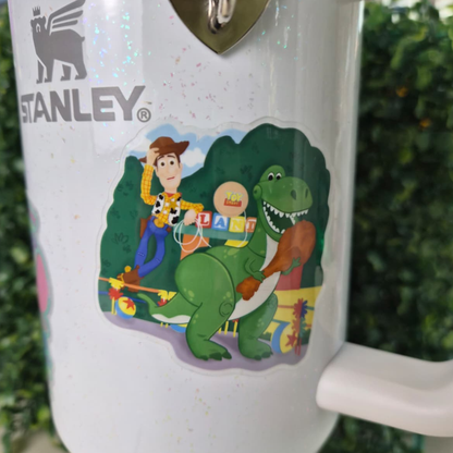 Stanley tumbler with cartoon design featuring a dinosaur and character, against a blurred green background.